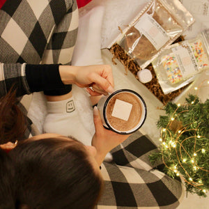 birds eye view of woman wearing pajamas and the logo socks, holding the logo mug with hot chocolate, and the cozy kit next to her