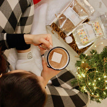 Load image into Gallery viewer, birds eye view of woman wearing pajamas and the logo socks, holding the logo mug with hot chocolate, and the cozy kit next to her