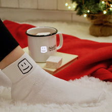 Load image into Gallery viewer, model&#39;s leg with logo white socks on, with logo mug with hot chocolate and marshmallow on top of a book