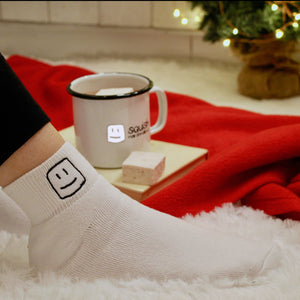 model's leg with logo white socks on, with logo mug with hot chocolate and marshmallow on top of a book